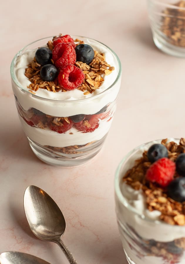 Layered summer berry breakfast parfait, with granola, in a glass cup on pink marble surface.