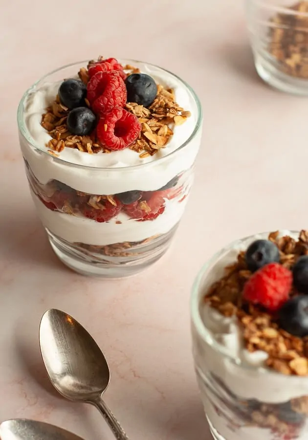 Layered summer berry breakfast parfait, with granola, in a glass cup on pink marble surface.