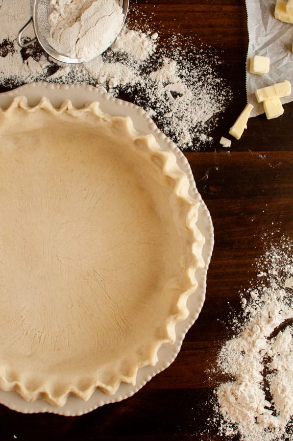 Prepared and crimped gluten free pie crust in ceramic dish surrounded by butter and flour on wooden surface
