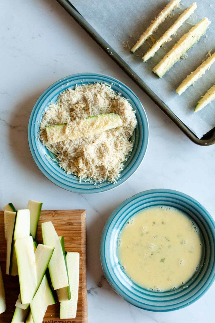 step photo for how to make zucchini fries, dip in egg mixture, dip in parmesan mixture and place on pan