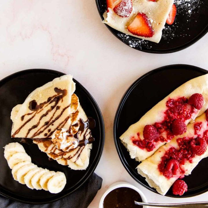 gluten free crepes served with fruit toppings on black plates sitting on a pink marble background