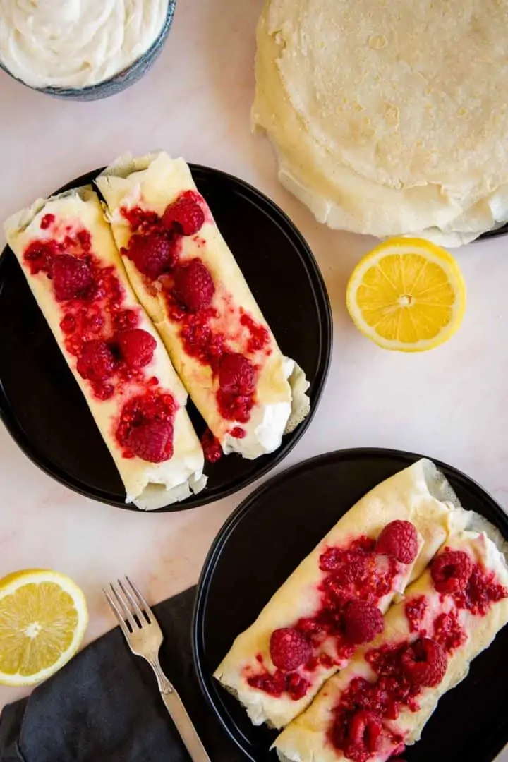 2 servings of ricotta cheese filled blintzes topped with fresh raspberries. Served on black plates on a pink marble surface