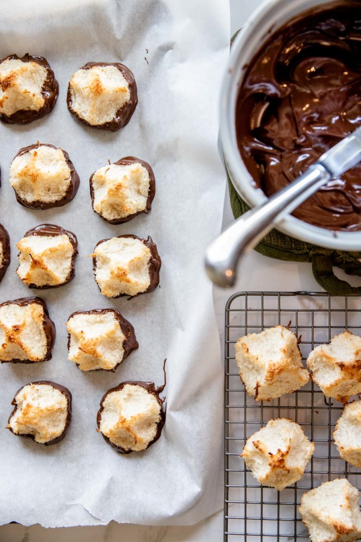 coconut macaroons being dipped in chocolate: dipped macaroons are on parchment and non-dipped macaroons are on a cooling rack sitting next to bowl of melted chocolate.