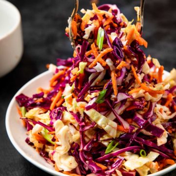 sweet and tangy coleslaw being tossed with serving utensils in a white bowl